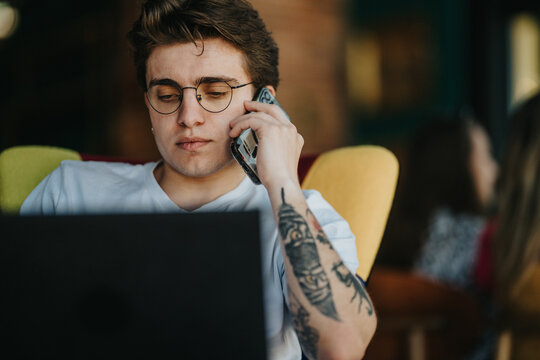 Two teenage friends work on a laptop while one talks on a smartphone. Casual cafe scene shows focused young people collaborating and communicating.