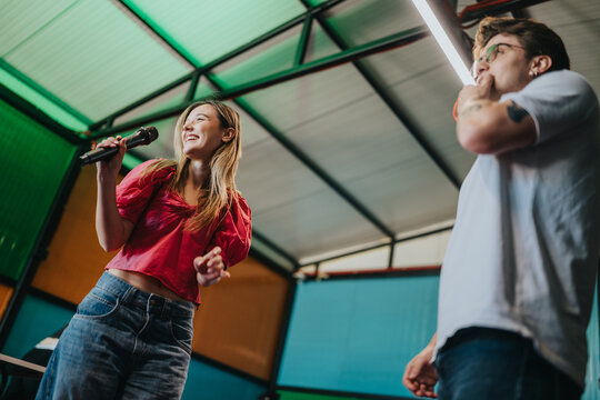 Two friends sing into a microphone while enjoying a casual rehearsal in a colorful indoor studio. The pair perform together, smiling and practicing music in a relaxed, modern space.