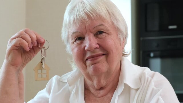 Happy senior woman holding key with house shaped keychain smiling at camera close up. Concept of real estate investment, mortgage and home loan