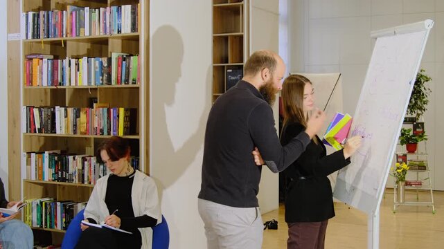 Teacher shows mistake in calculation to woman student on whiteboard in library. Mature tutor explains lesson to teenagers in classroom