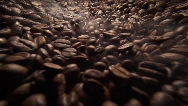Wide angle sliding shot of roast coffee beans with gentle smoke on a dark background. Fresh coffee beans roasting with aromatic steam slow motion video 120 fps. 