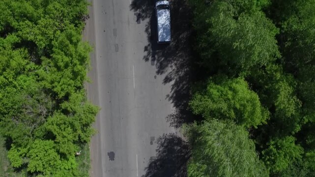 Top-down drone shot following a white van as it drives along an asphalt road with light traffic, flanked by lush green trees on a bright day.

