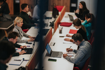 Business professionals collaborate at a long shared desk, using laptops and taking notes. A modern...
