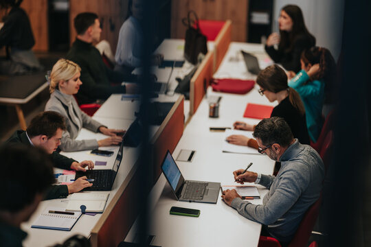 Business professionals collaborate at a long shared desk, using laptops and taking notes. A modern coworking office scene showing focused colleagues working side by side.