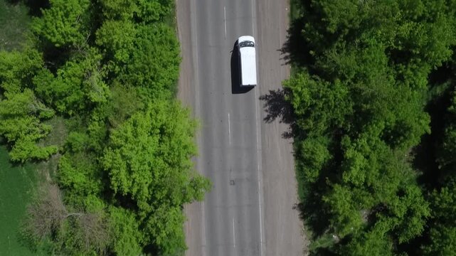 Top-down drone shot following a white van as it drives along an asphalt road with light traffic, flanked by lush green trees on a bright day.

