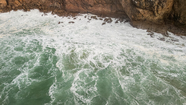 Aerial view of rough turquoise waves crashing against the rugged limestone cliffs with white sea foam Sagres, Faro, Portugal.