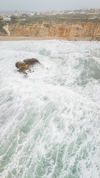 Aerial view of rough Atlantic waves crashing against a rock formation and steep cliffs near the town of Sagres, Faro, Portugal.