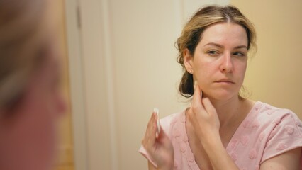 Woman in bathroom gently applying skincare with a cotton pad while looking in mirror, focused on morning facial cleansing and moisturizing routine for healthy, glowing skin © MIKHAIL