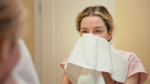 Young woman drying her face with a soft white towel in a bathroom mirror reflection, enjoying a fresh morning skincare and self care routine at home