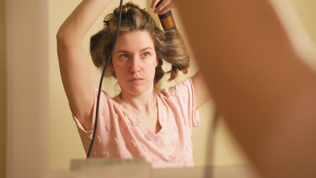 Young woman styling blonde hair in front of bathroom mirror, carefully curling strands with an electric styler as part of her morning self care and beauty routine