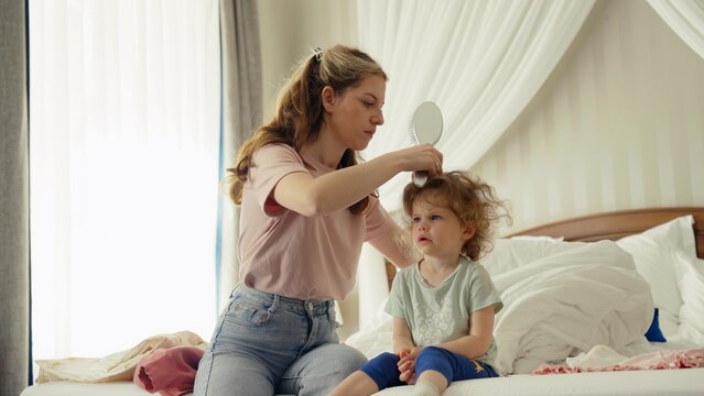 Mother carefully brushing her young daughter's messy hair while sitting on the bed during their daily morning routine, nurturing a moment of family bonding and care