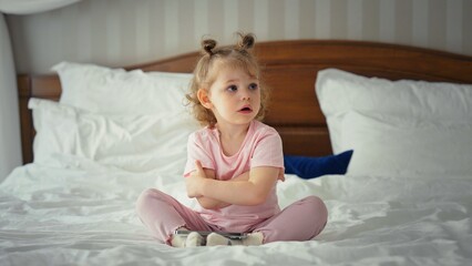 Fototapeta premium Little girl with pigtails wearing pajamas and sitting cross legged on a white bed, expressing feelings of disappointment, sadness, and grumpiness with crossed arms