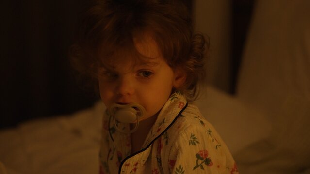 Toddler girl in floral pajamas with a pacifier, sitting in a dim bedroom ready for sleep, sleepy eyes and calm expression conveying comfort, routine, and gentle bedtime warmth