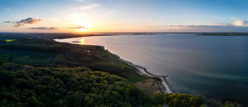 Aerial view of Bockholmwik coastline with lush green forests and calm waters under a golden sunset sky in Munkbrarup, Schleswig-Holstein, Germany.