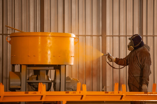 Professional industrial painter worker wearing safety respirator mask painting heavy metal machinery with spray gun in manufacturing factory. Commercial business production and occupational coating.