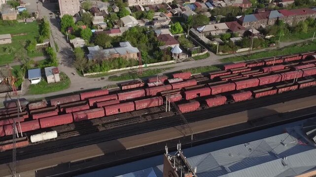 High angle aerial drone view of a railway station with red cargo wagons parked on tracks, surrounded by residential buildings in a small town during sunny spring day.

