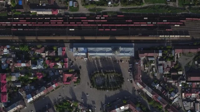 High angle aerial drone view of a railway station with red cargo wagons parked on tracks, surrounded by residential buildings in a small town during sunny spring day.

