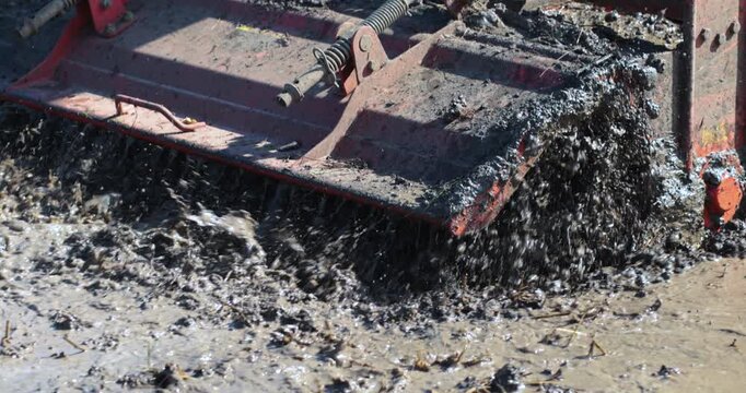 Tractor tilling muddy rice field with splashing water and soil. Agricultural machinery working in wet farmland, ideal for farming, rural life, and cultivation concepts, close-up