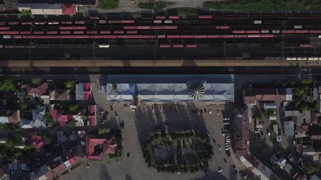 High angle aerial drone view of a railway station with red cargo wagons parked on tracks, surrounded by residential buildings in a small town during sunny spring day.

