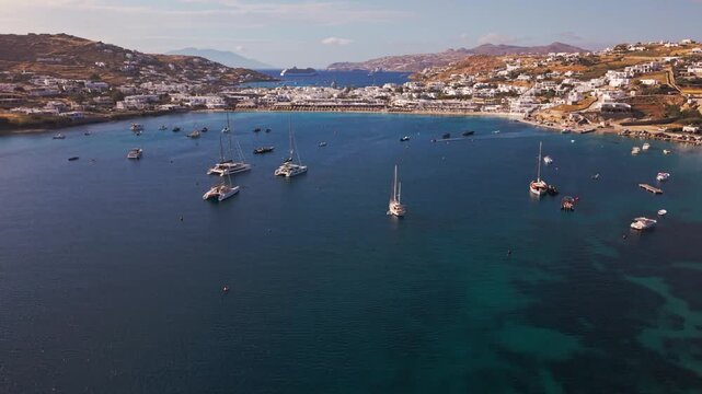 High angle panoramic drone footage of Ornos beach and bay in Mykonos island, Greece. Luxury yachts and boats anchored in the turquoise Aegean sea surrounded by traditional whitewashed villas.