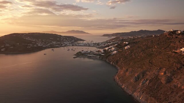 High angle panoramic drone footage of golden hour sunset over Ornos bay in Mykonos island, Greece. Luxury yachts and boats anchored on calm sea with sun reflection and coastal villas background.