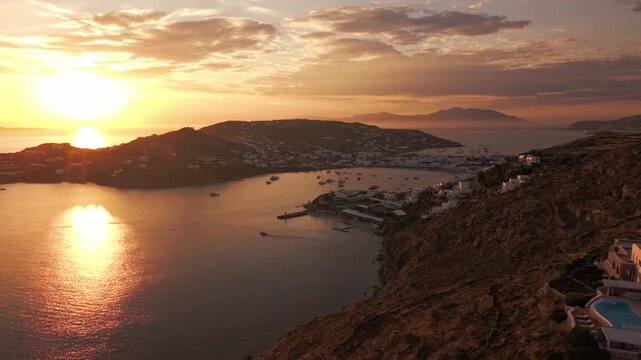 High angle panoramic drone footage of golden hour sunset over Ornos bay in Mykonos island, Greece. Luxury yachts and boats anchored on calm sea with sun reflection and coastal villas background.