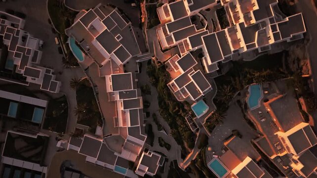 Birds-eye view drone footage of luxury hotel resort architecture with swimming pools at sunset. Geometric pattern of white villas and swimming pools during golden hour in Mykonos, Greece.