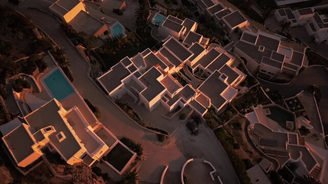 Birds-eye view drone footage of luxury hotel resort architecture with swimming pools at sunset. Geometric pattern of white villas and swimming pools during golden hour in Mykonos, Greece.