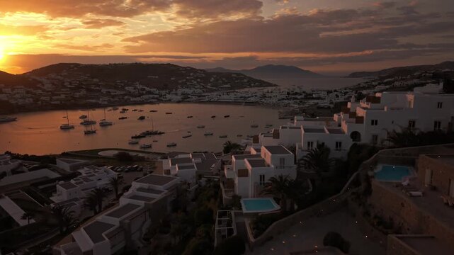 High angle panoramic drone footage of golden hour sunset over Ornos bay in Mykonos island, Greece. Luxury yachts and boats anchored on calm sea with sun reflection and coastal villas background.