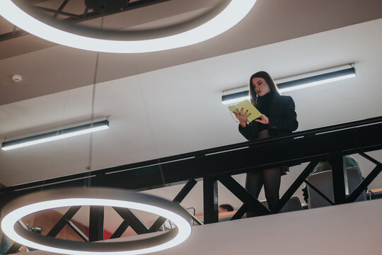 A businesswoman reads a notebook while standing on a mezzanine balcony in a modern office. She studies notes under large circular lights overlooking the workspace.