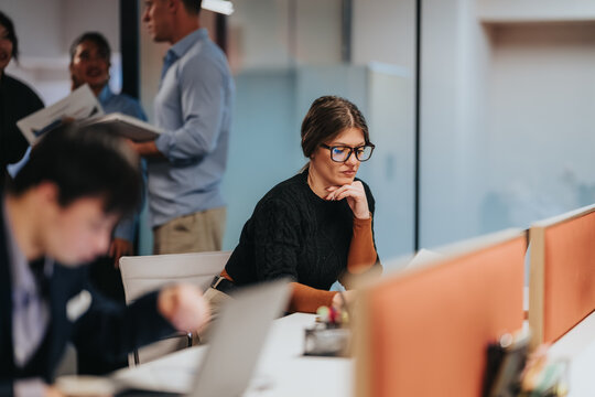 A businesswoman wearing glasses concentrates at her desk while working on documents. Colleagues collaborate and move through the blurred open-plan office behind her.
