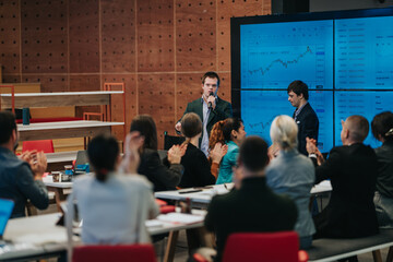 Obraz premium A business presenter shows financial charts on large screens while colleagues applaud during a meeting. The corporate team listens and reacts to live market data displayed on monitors.
