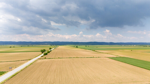 Aerial view of vast agricultural fields and a straight road under a dramatic cloudy sky in Novi Sad, Vojvodina, Serbia.