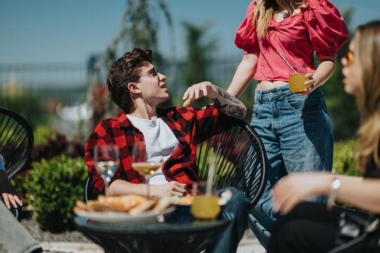 A group of teenage friends relax outdoors as a teen in a red plaid shirt talks while seated. A standing friend holds a drink during a casual summer patio gathering.