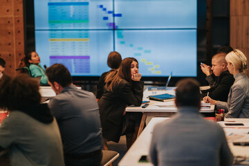 Obraz premium A business team sits around a table during a project meeting with a large screen presentation. Colleagues listen, take notes and appear thoughtful while reviewing project timelines.