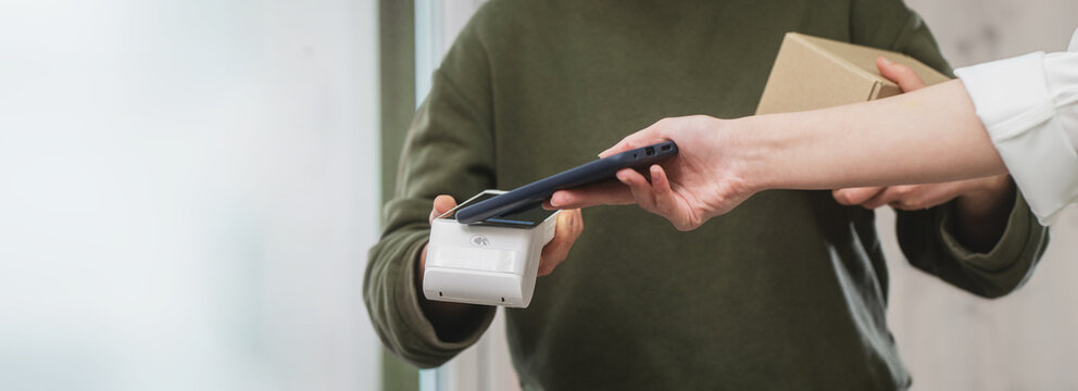 Woman paying with smartphone for parcel delivery for courier. Electronic cashless payments. Online shopping. A woman enters a PIN code on a wireless payment terminal.