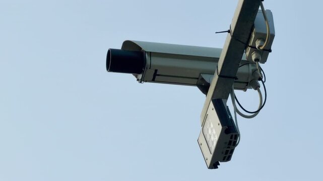 An automated surveillance camera is fixed onto a diagonal metal beam. The device aims forward to monitor traffic from above. It stands out against a pale blue sky.