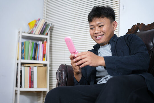 A young Asian man relaxes on a sofa reading text messages on a pink flip phone while smiling. Using a flip phone for communication.
