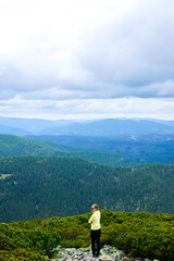 Fototapeta premium Beautiful mountains and sitting young woman on mountain peak in summer. Landscape with girl in yellow on the mountain peak, forest, hills , blue sky with clouds and sunbeams. Travel, hiking concept