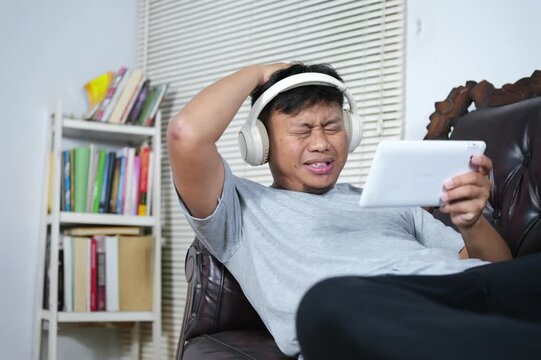 An Asian man is focused on playing a game on his smartphone, lying on the couch with a sad and disappointed expression after losing. He's enjoying the weekend by relaxing and playing games.
