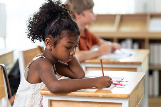Young schoolgirl writes assignment with pencil at wooden desk during elementary classwork, practicing responsibility, attention skill, study habit, independent learning while classmates work nearby.