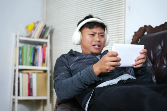 An Asian man is focused on playing a game on his smartphone, lying on the couch with a sad and disappointed expression after losing. He's enjoying the weekend by relaxing and playing games.
