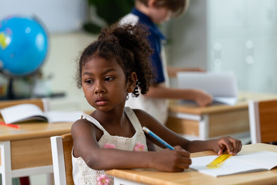 Elementary girl studies notebook with pencil and ruler at desk while classmate works behind, back to school classroom routine supports writing practice, attention, confidence, study discipline.