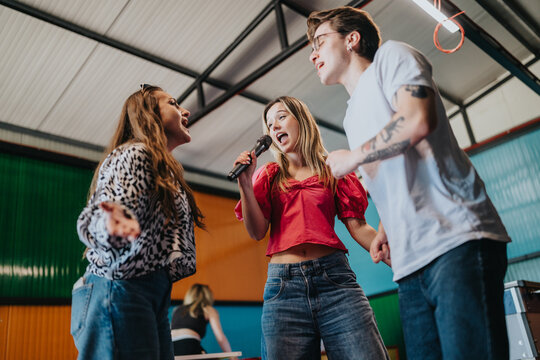 Three friends sing into a microphone while enjoying a casual indoor hangout. They perform together, laughing and having fun in a colorful community space.