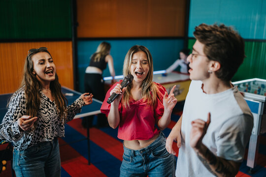 A group of young friends singing with a microphone in a colorful game room. They are friends enjoying karaoke, dancing and laughing together near table games.