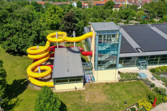 Aerial view of Konigsbad Forchheim featuring vibrant yellow and red water slides and a modern glass staircase tower surrounded by lush green trees in Forchheim, Bayern, Germany.