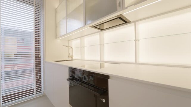 Cabinet LED backlight turns on inside a white kitchen illuminating empty countertop with built-in induction stove, black oven and faucet for house interior design or apartment renovation.
