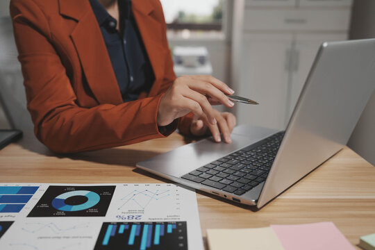 Business professional in rust blazer using laptop at desk, holding pen, with printed charts and graphs in foreground; modern office setting, shallow depth of field.