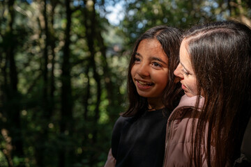 Naklejka premium A mother and daughter smile sweetly in a sun-drenched wood. Sunbeams filter through the tall trees behind them. One of them looks towards the camera with a warm expression. The other looks on tenderly
