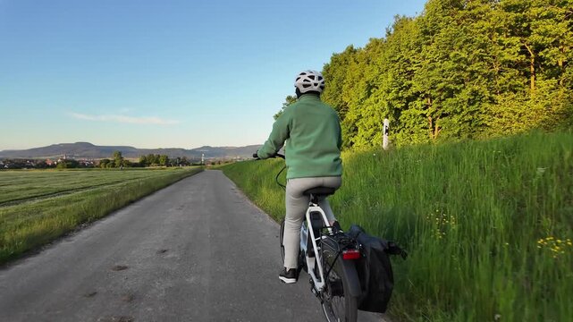 Woman cyclist riding electric bicycle on rural road in evening countryside  